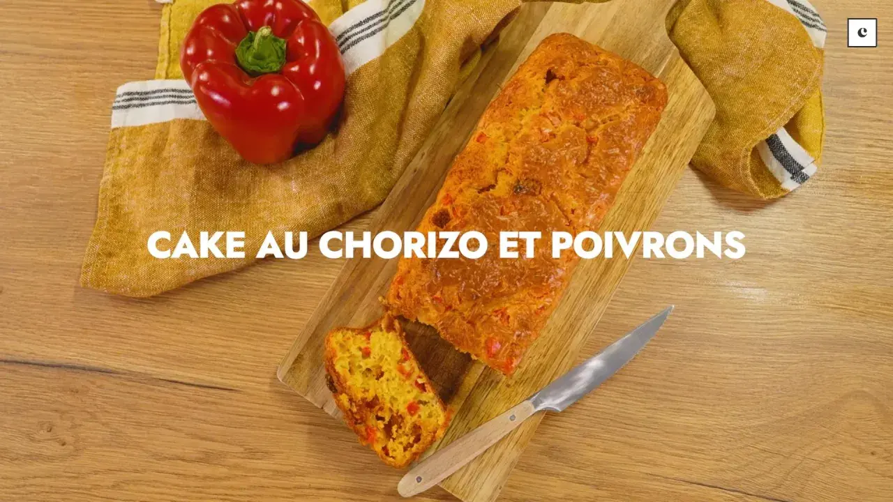 Un cake au chorizo et poivrons, coupé en tranche, sur une planche en bois. Un poivron rouge est posé à côté.