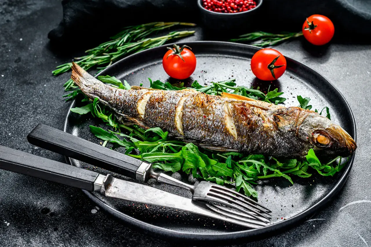 Bar au barbecue, entier et doré, servi sur un lit de roquette avec des tomates cerises et des herbes. Un délice à savourer !