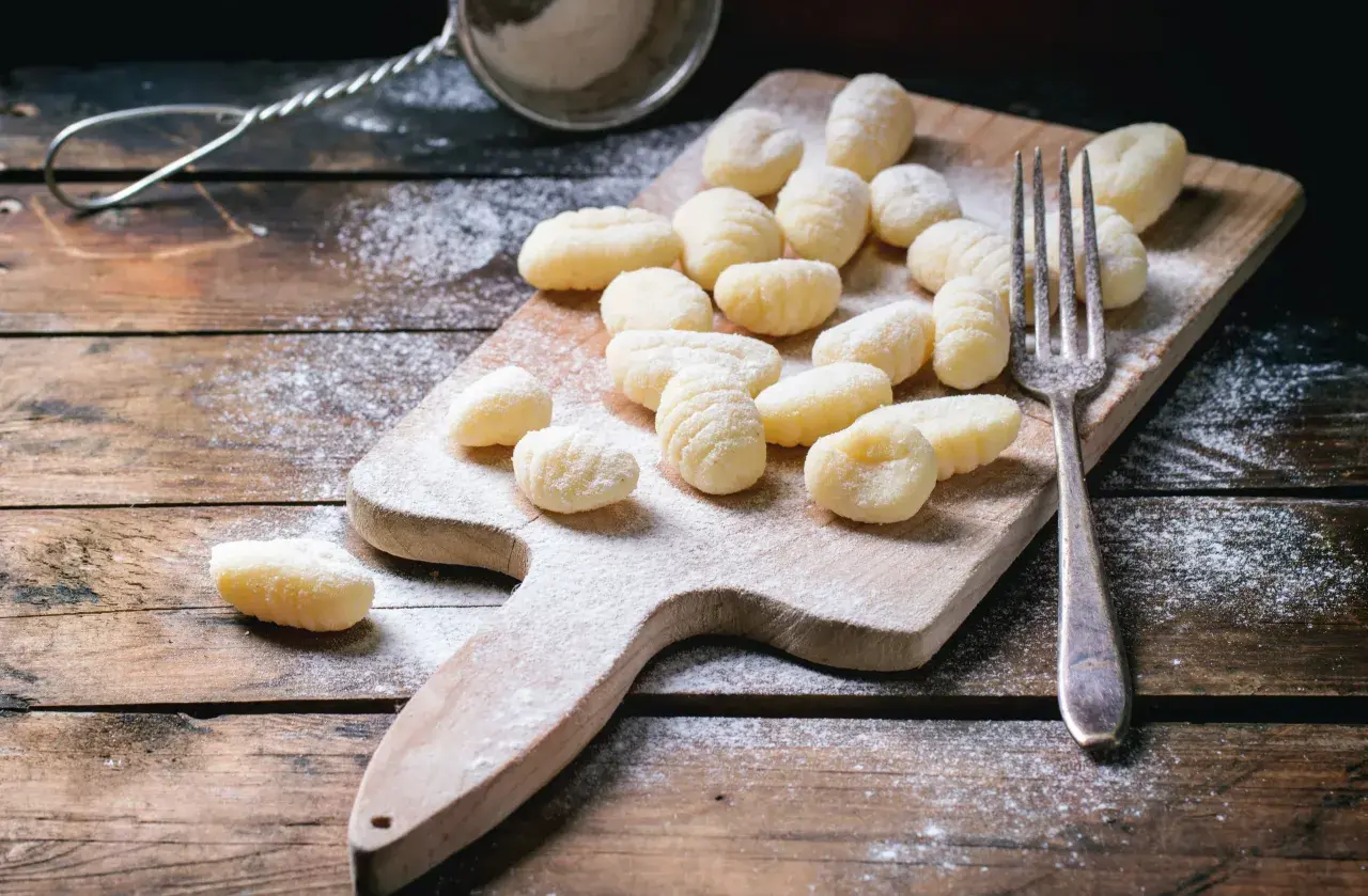 Gnocchis faits maison, prêts pour la cuisson. Une fourchette et une passoire à farine sur une planche en bois saupoudrée de farine, parfaits pour une recette de gnocchi.