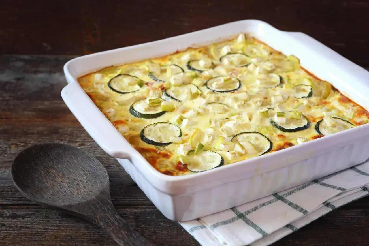 Un plat blanc rectangulaire contient un gratin de courgettes moelleux, garni de rondelles de courgettes et de morceaux de fromage. Une cuillère en bois repose à côté.