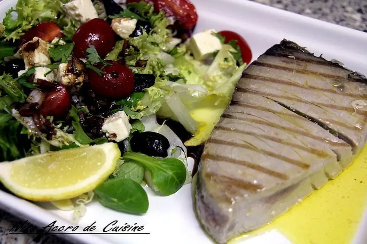 Un steak de thon au barbecue, grillé à la perfection, servi avec une salade fraîche composée de laitue, tomates cerises, olives noires, feta et un quartier de citron.