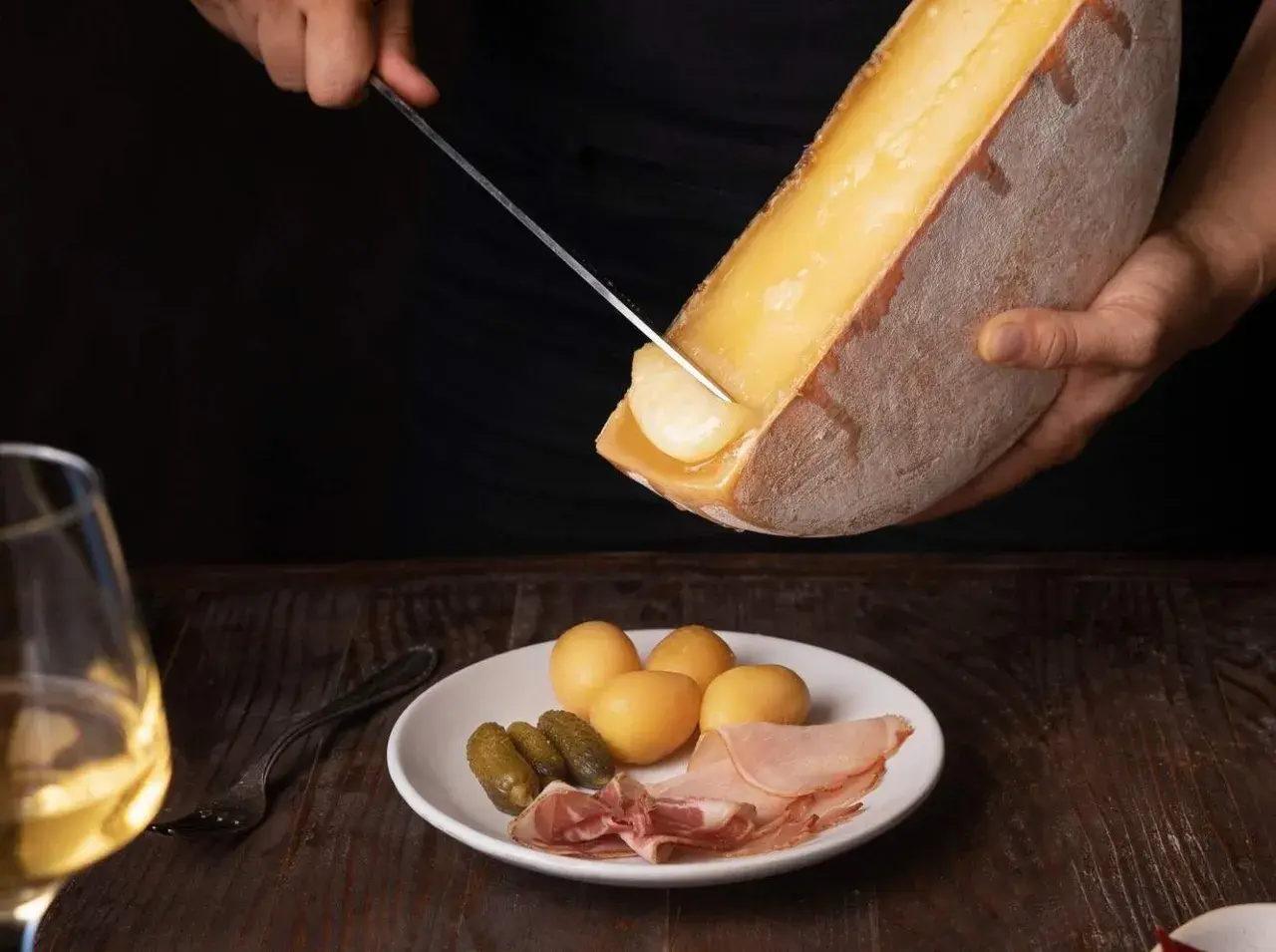 Un verre de vin blanc, parfait pour accompagner la raclette. Sur l'assiette, pommes de terre, cornichons et charcuterie.