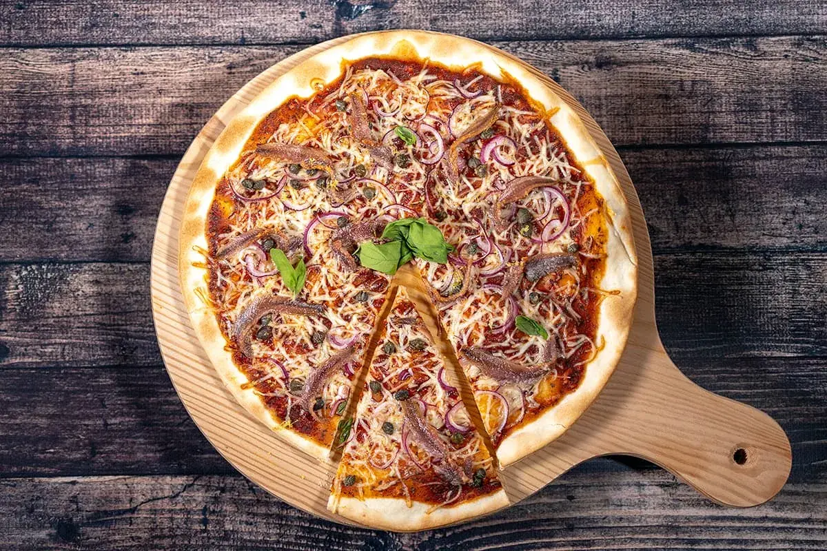 Une pizza savoureuse aux anchois, oignons rouges et câpres, garnie de fromage râpé et de basilic frais, prête à être dégustée.