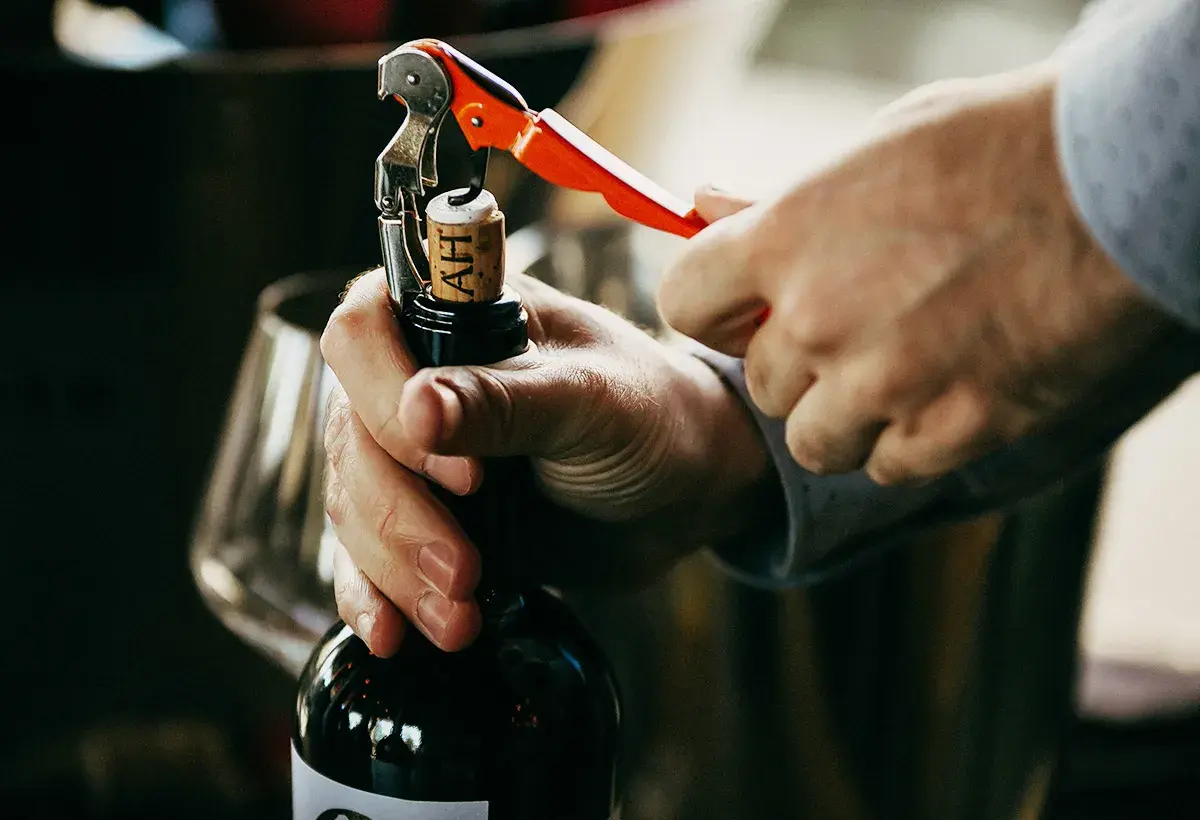 Un homme ouvre une bouteille de vin rouge avec un tire-bouchon. La scène évoque le plaisir de la dégustation et la conservation du vin.