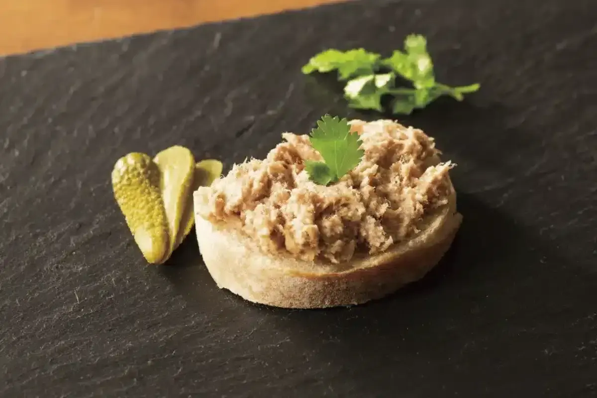 Tranche de pain garnie de rillettes de porc, accompagnée de cornichons et d'une feuille de persil.