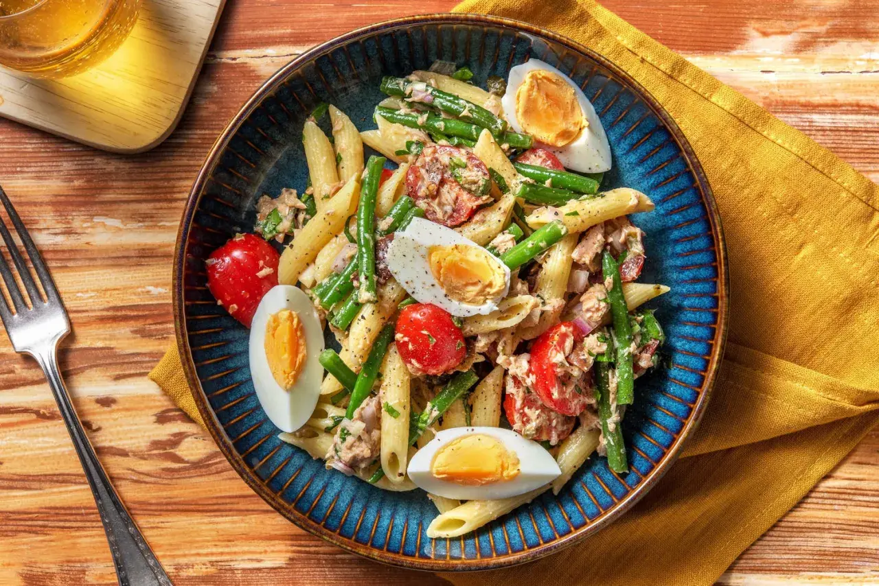 Salade de penne colorée avec haricots verts, tomates cerises, thon et œufs durs, servie dans un plat bleu.