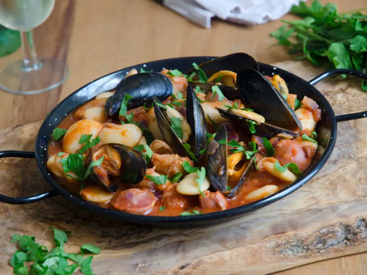 Un plat de moules au chorizo, garni de persil frais, servi dans une poêle noire sur une planche en bois.