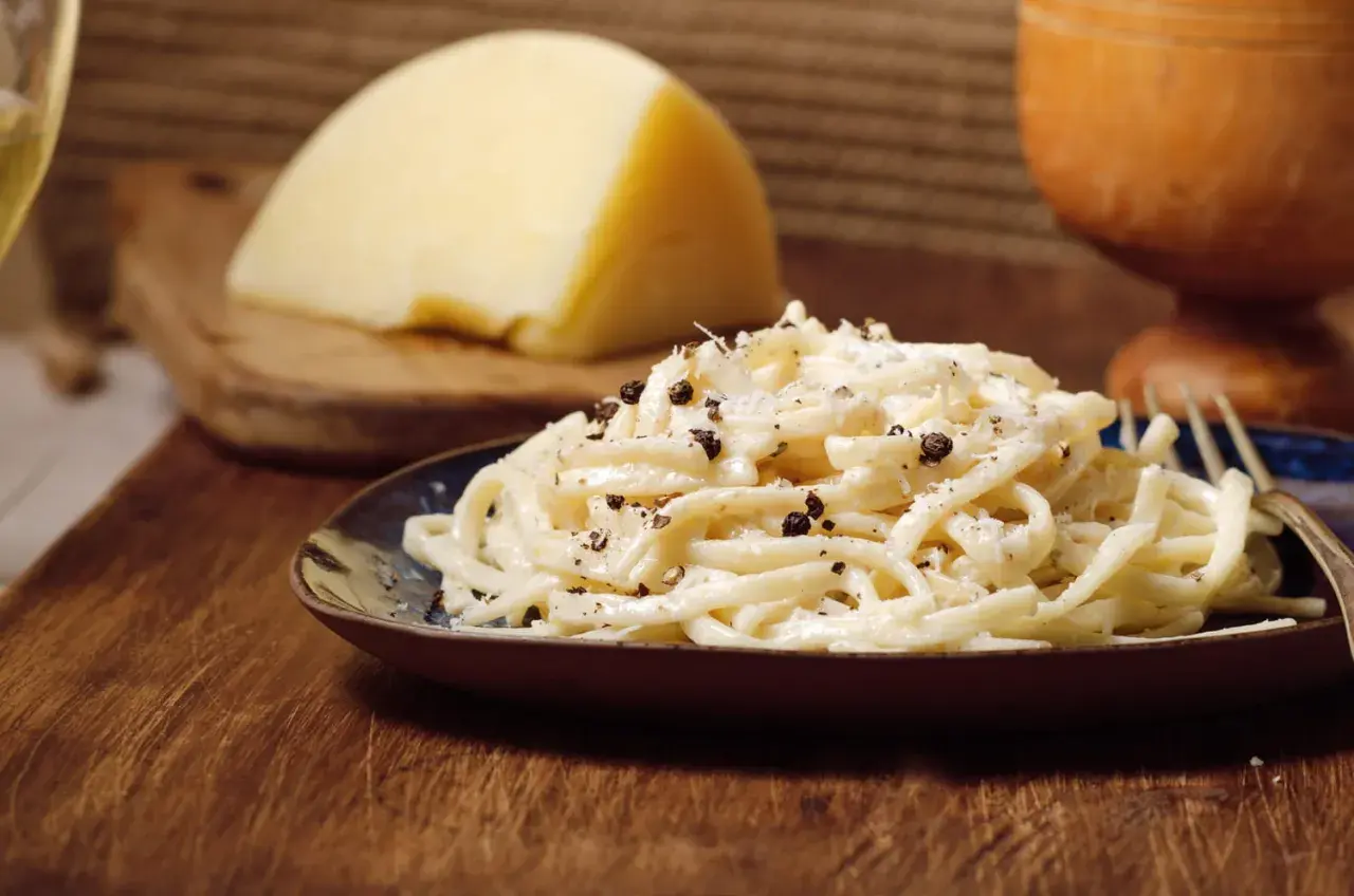 Un plat de pâtes crémeux, garni de poivre noir concassé, évoque le goût authentique du cacio e pepe. Un morceau de fromage pecorino est visible en arrière-plan.