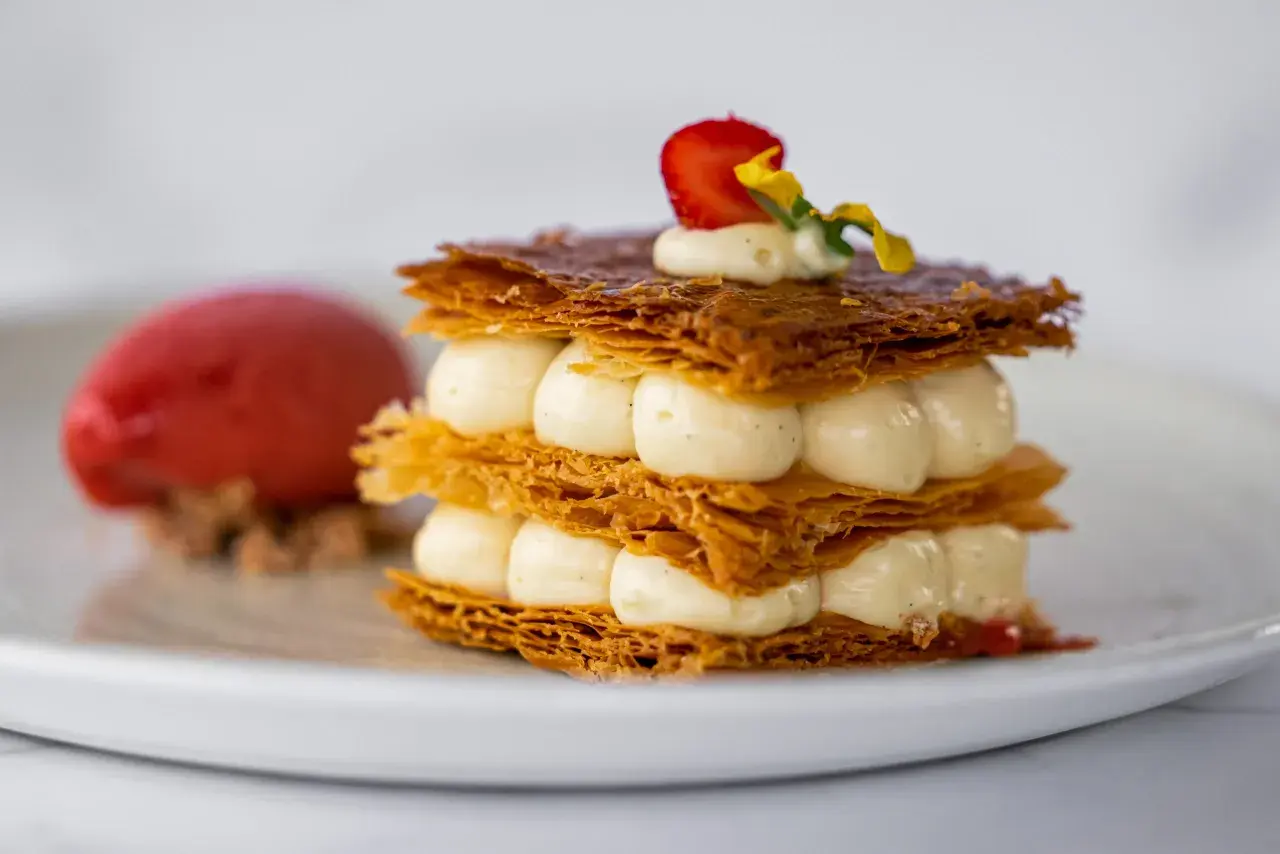 Un délicieux millefeuille croustillant, garni de crème onctueuse et d'une fraise, servi avec une boule de sorbet rouge.