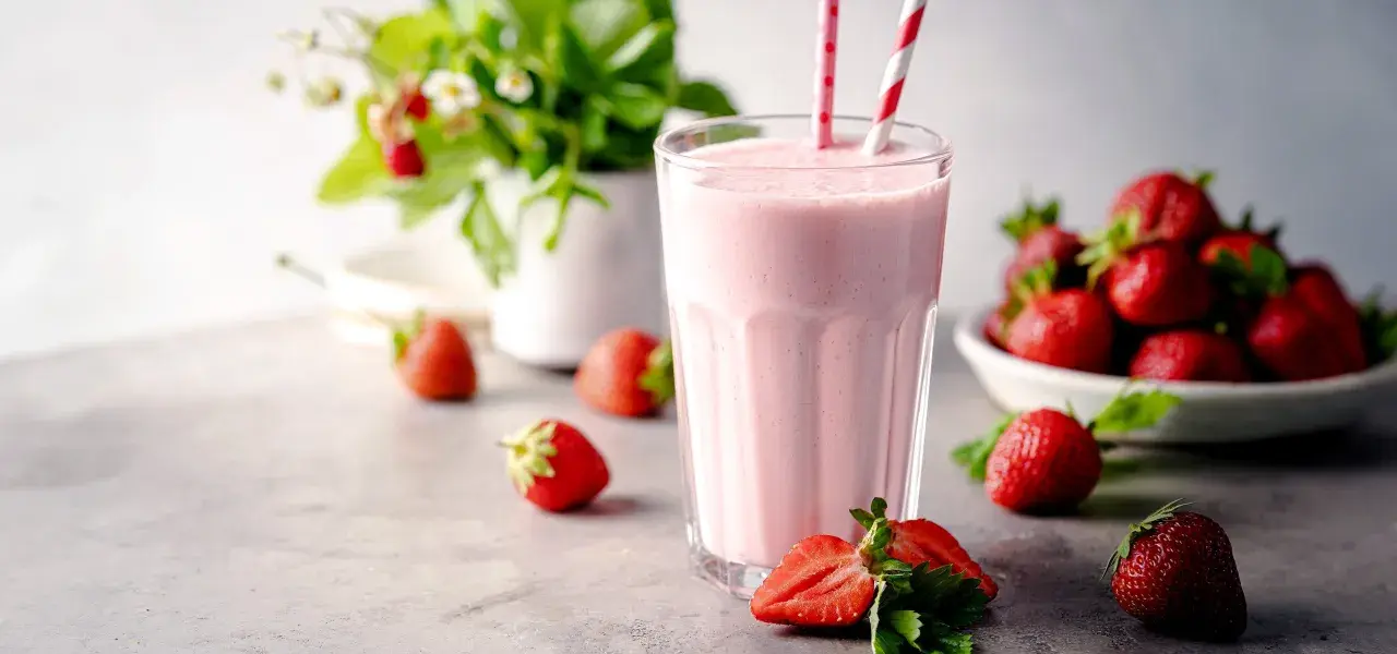 Un délicieux milkshake fraise crémeux, garni de pailles rayées, entouré de fraises fraîches et d'un plant de fraises en arrière-plan.