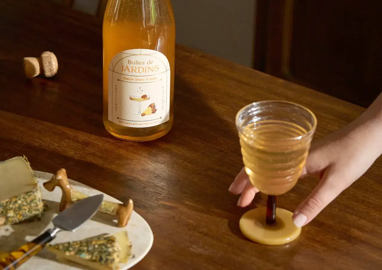 Une main saisit un verre de ce qui pourrait être le meilleur vin blanc moelleux, à côté d'une bouteille "Bulles de Jardins" et de fromages.