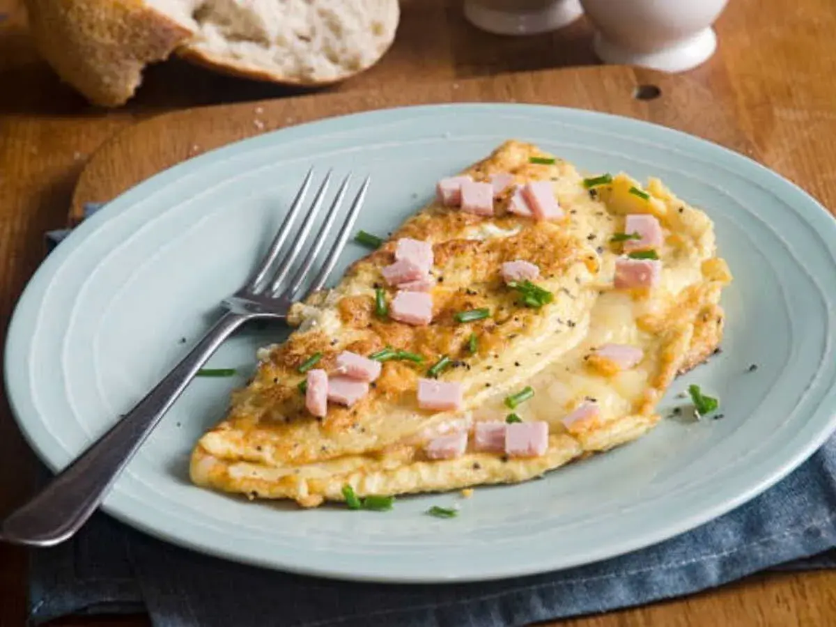 Une délicieuse omelette jambon fromage, garnie de ciboulette et de poivre noir, prête à être dégustée avec une tranche de pain croustillant.