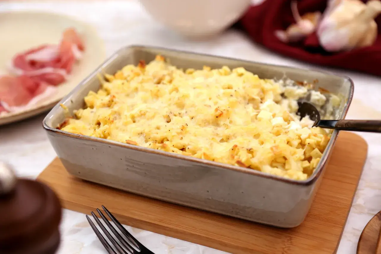 Un plat de gratin de crozet doré et gratiné, prêt à être dégusté. Une assiette de jambon cru est visible à gauche.