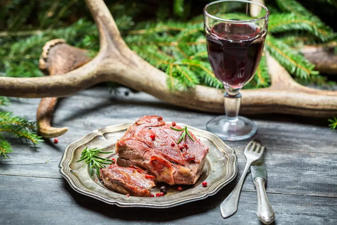 Un plat de chevreuil rôti, garni de romarin et de baies roses, accompagné d'un verre de vin rouge. La question est : quel vin avec du chevreuil ?
