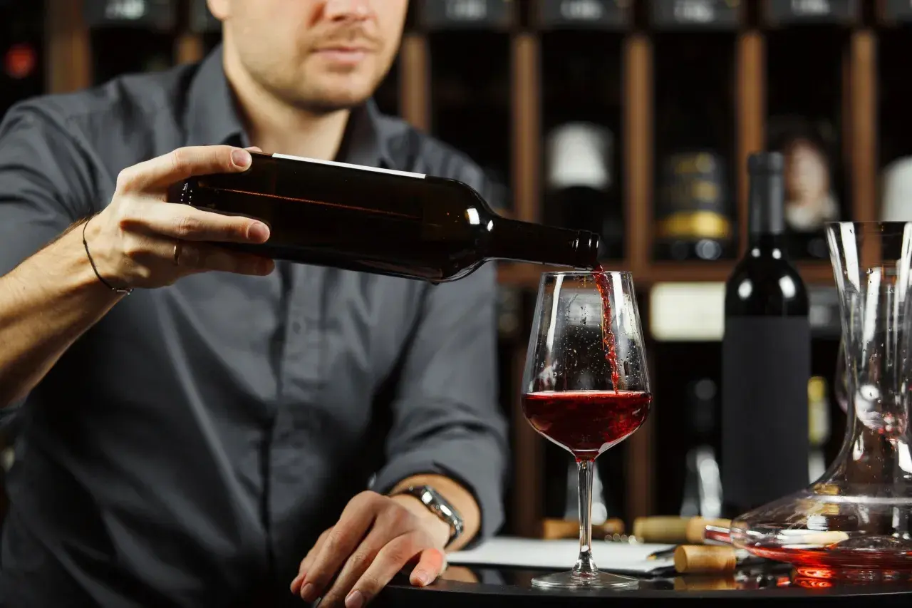 Un homme verse un vin rouge profond dans un verre, une cave à vin en arrière-plan. Quel vin rouge savoureux !