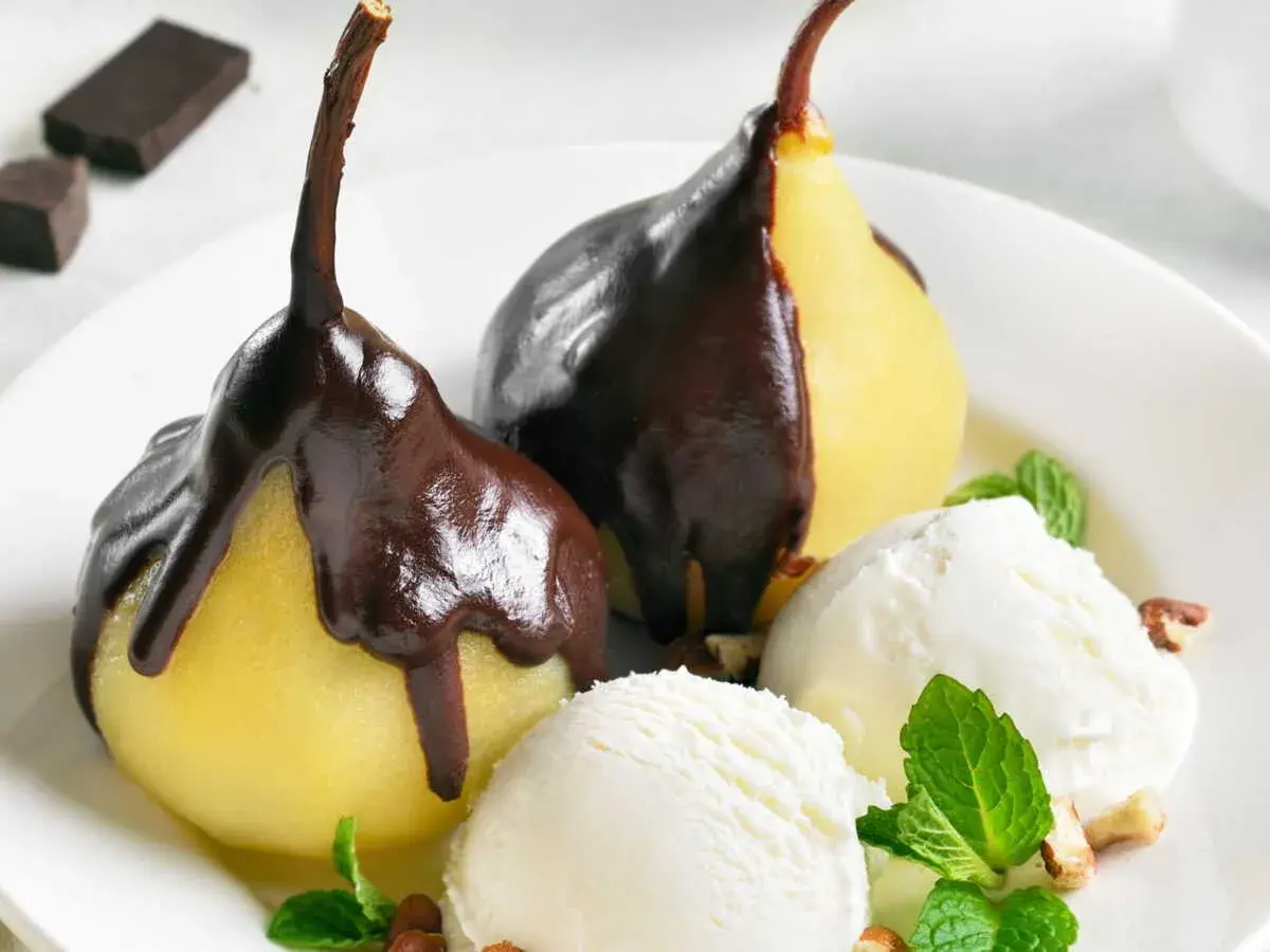 Poires pochées nappées de chocolat noir, servies avec de la glace à la vanille et des éclats de noisettes. Un délice de poire Belle Hélène.