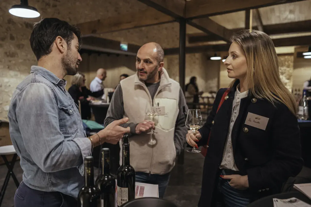 Trois personnes discutent autour d'une table de dégustation. L'une d'elles tient un verre de vin, peut-être un rosé de la terre du midi.