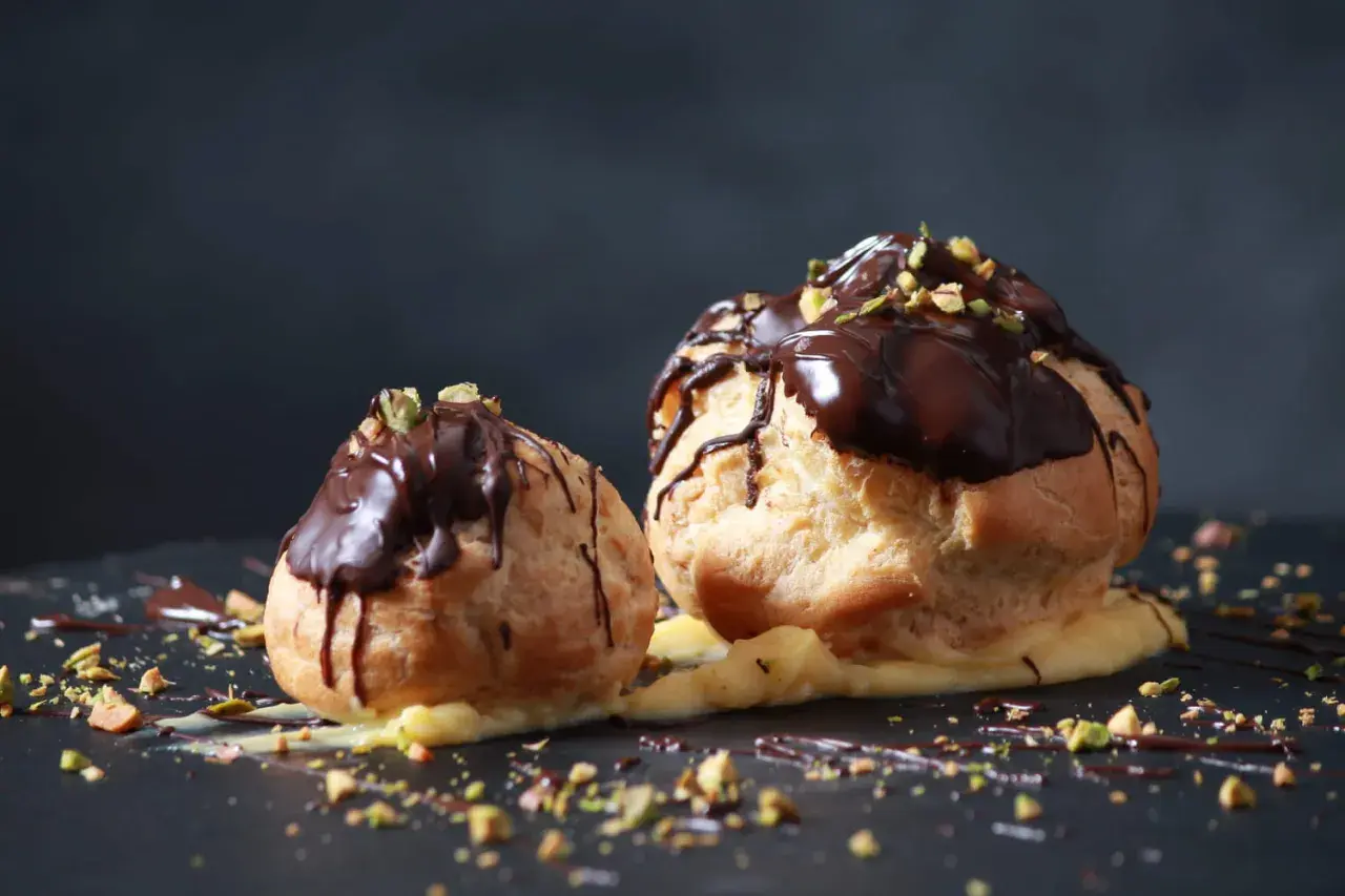 Deux profiteroles au chocolat, garnis de crème pâtissière et saupoudrés de pistaches concassées, sur un fond sombre.