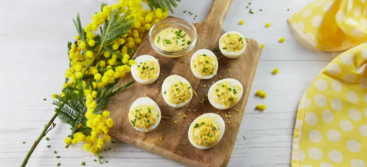 Des œufs mimosa garnis d'une crème onctueuse et de ciboulette, disposés sur une planche en bois, avec des fleurs de mimosa en arrière-plan.