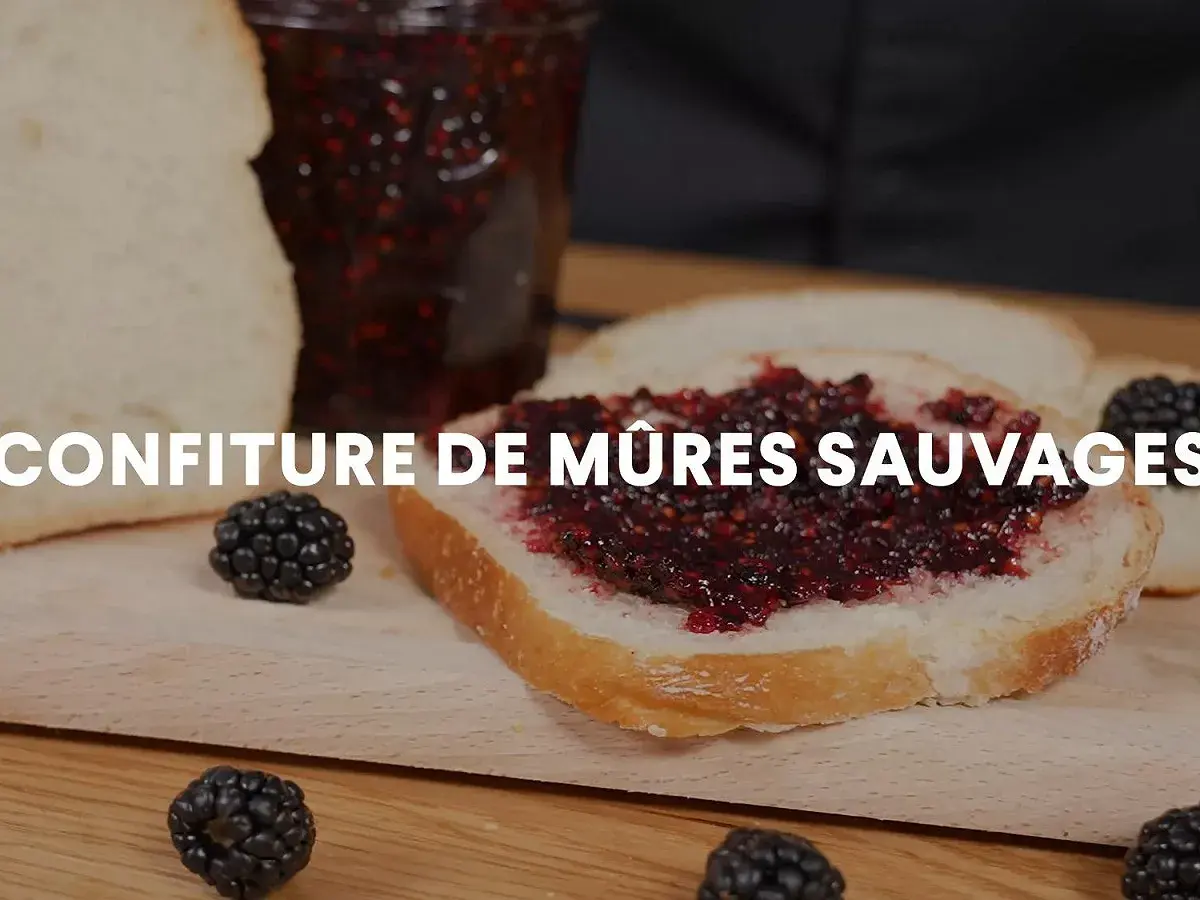 Tranche de pain tartinée de confiture de mûres sauvages, avec des mûres fraîches et un pot de confiture en arrière-plan.