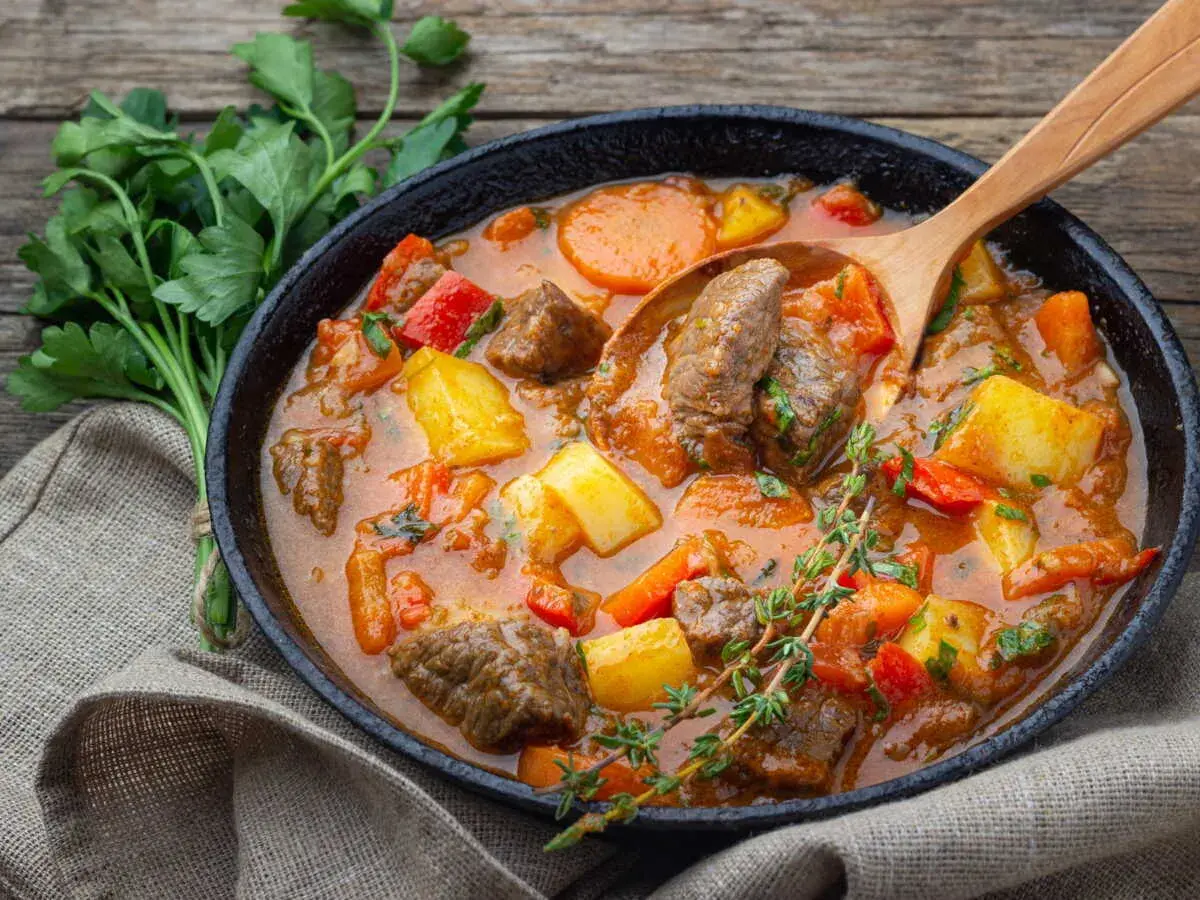 Un plat fumant de recette goulash hongroise traditionnelle, avec des morceaux de bœuf, pommes de terre, carottes et poivrons rouges, servi dans une poêle en fonte avec une cuillère en bois.