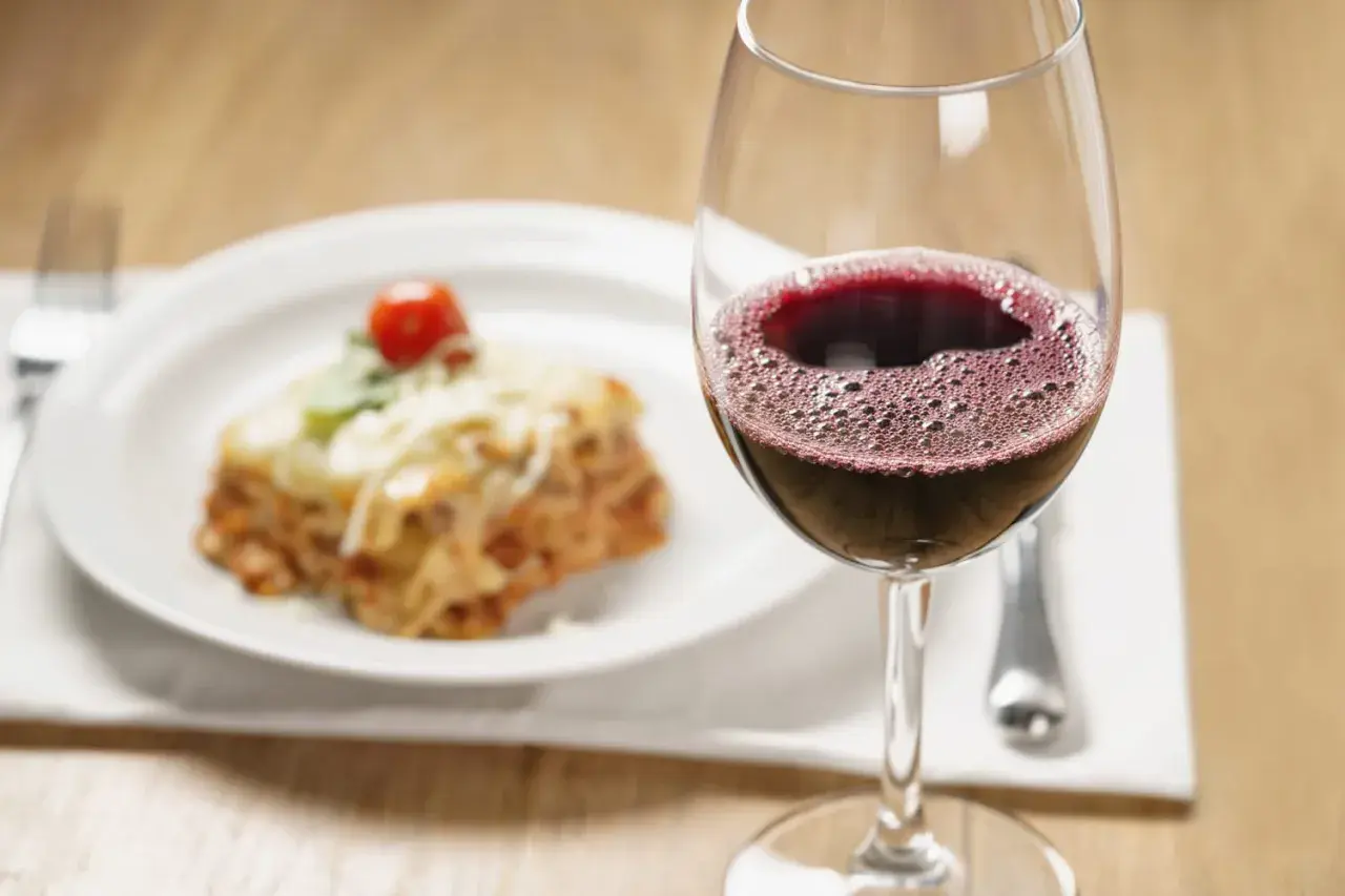 Un verre de vin rouge pétillant à côté d'une assiette de lasagnes au fromage fondu. La question est : quel vin avec des lasagnes ?