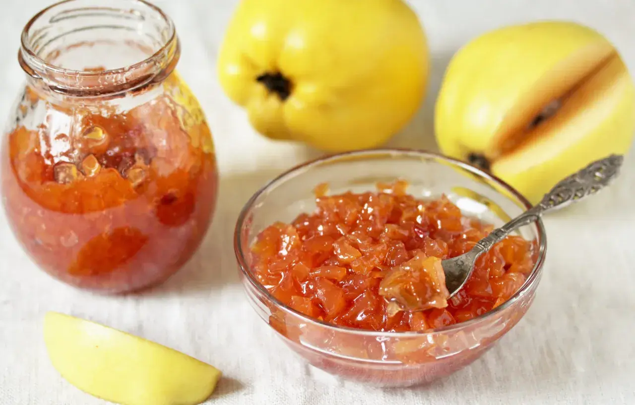 Confiture de coings sans les peler, servie dans un bocal et un bol, avec des coings entiers et une tranche en arrière-plan.