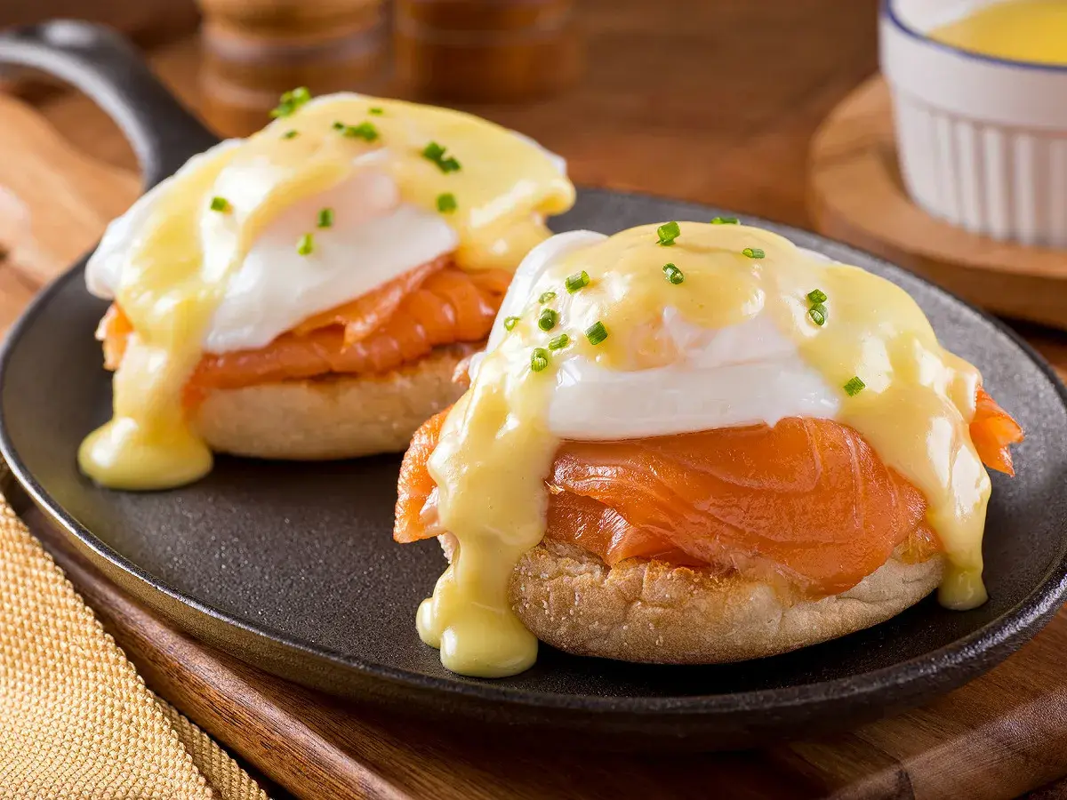 Deux oeufs Bénédicte au saumon fumé, nappés de sauce hollandaise et parsemés de ciboulette, servis sur un muffin anglais dans une poêle.