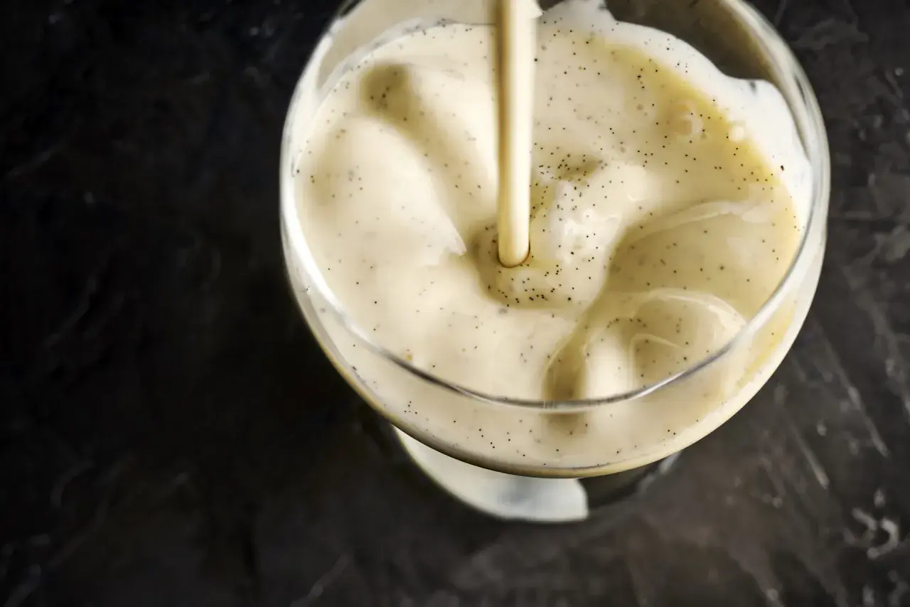 Une crème anglaise onctueuse, parsemée de points de vanille, est versée dans un verre.