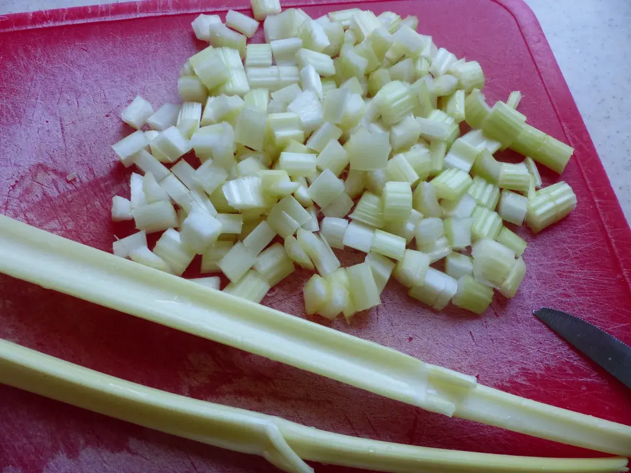 Céleri branche coupé en dés sur une planche rouge, prêt pour une recette. Deux branches entières sont posées devant.