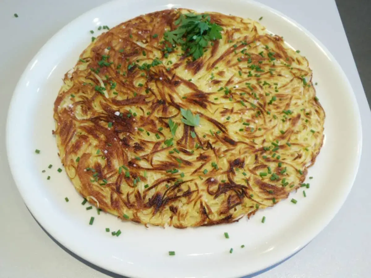 Une pomme darphin dorée et croustillante, garnie de ciboulette fraîche et de persil, servie sur une assiette blanche.