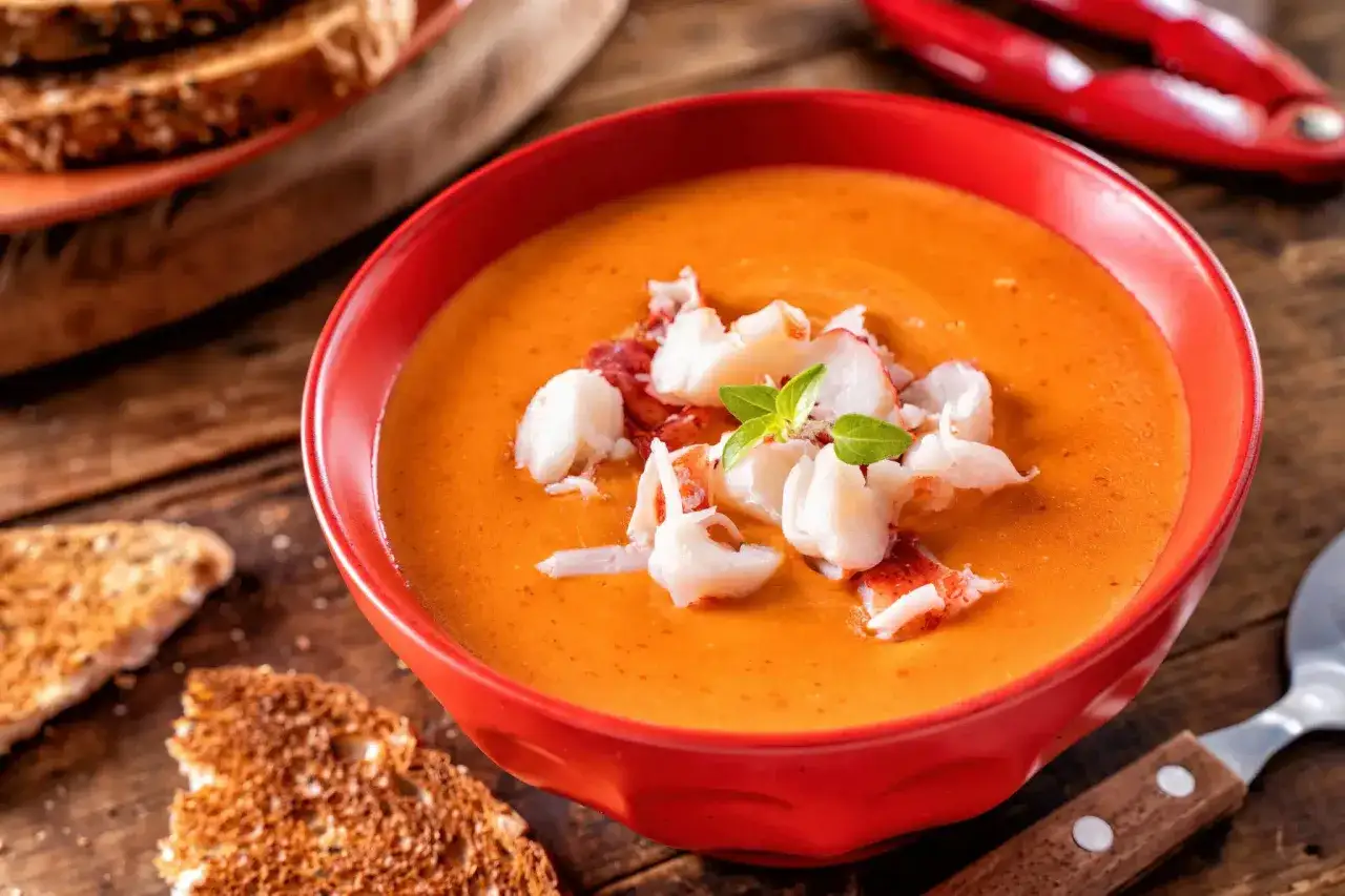 Un bol rouge vif de bisque de homard, garni de morceaux de homard et de basilic. Des toasts grill&eacute;s et un casse-noix de homard rouge sont visibles en arri&egrave;re-plan.
