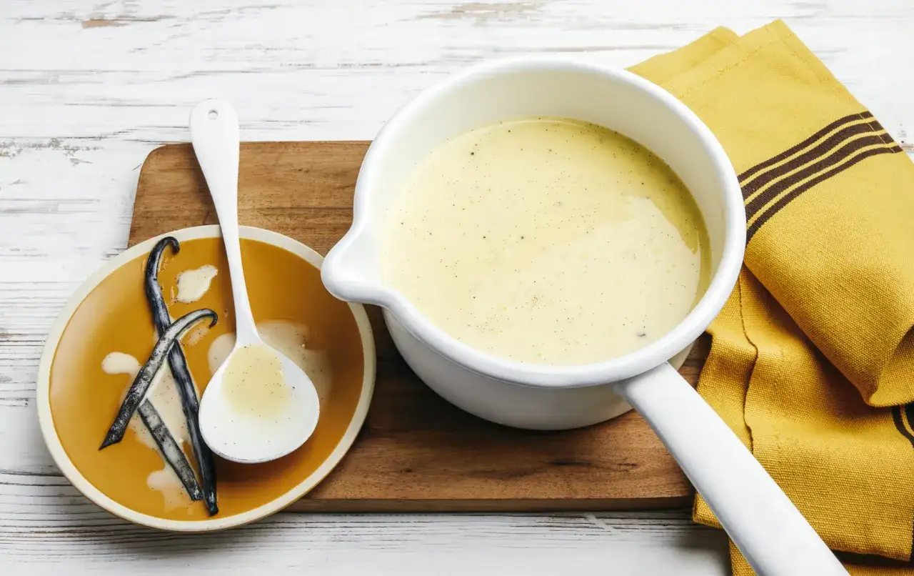 Une casserole de crème anglaise onctueuse, des gousses de vanille sur une assiette orange, et une cuillère remplie de cette délicieuse crème.