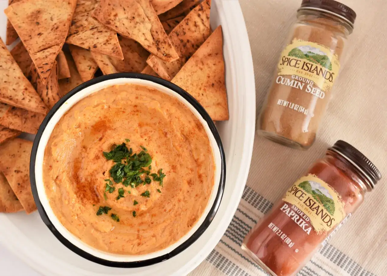 Un bol de purée de pois chiche garni de persil, servi avec des chips pita croustillantes. Des pots d'épices, cumin et paprika, sont à côté.