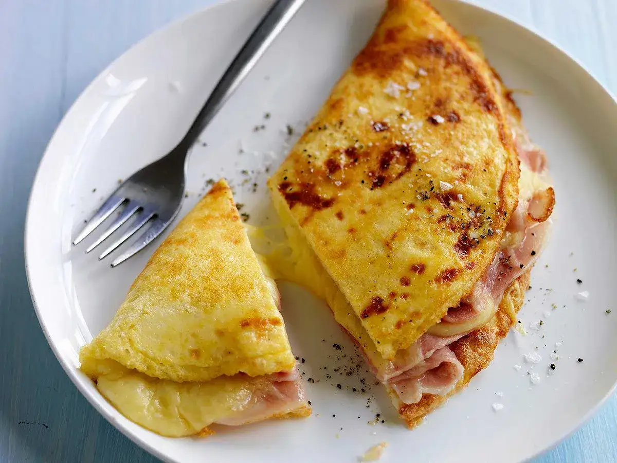 Une omelette jambon fromage pliée en deux, garnie de jambon et de fromage fondant, servie sur une assiette blanche avec une fourchette.