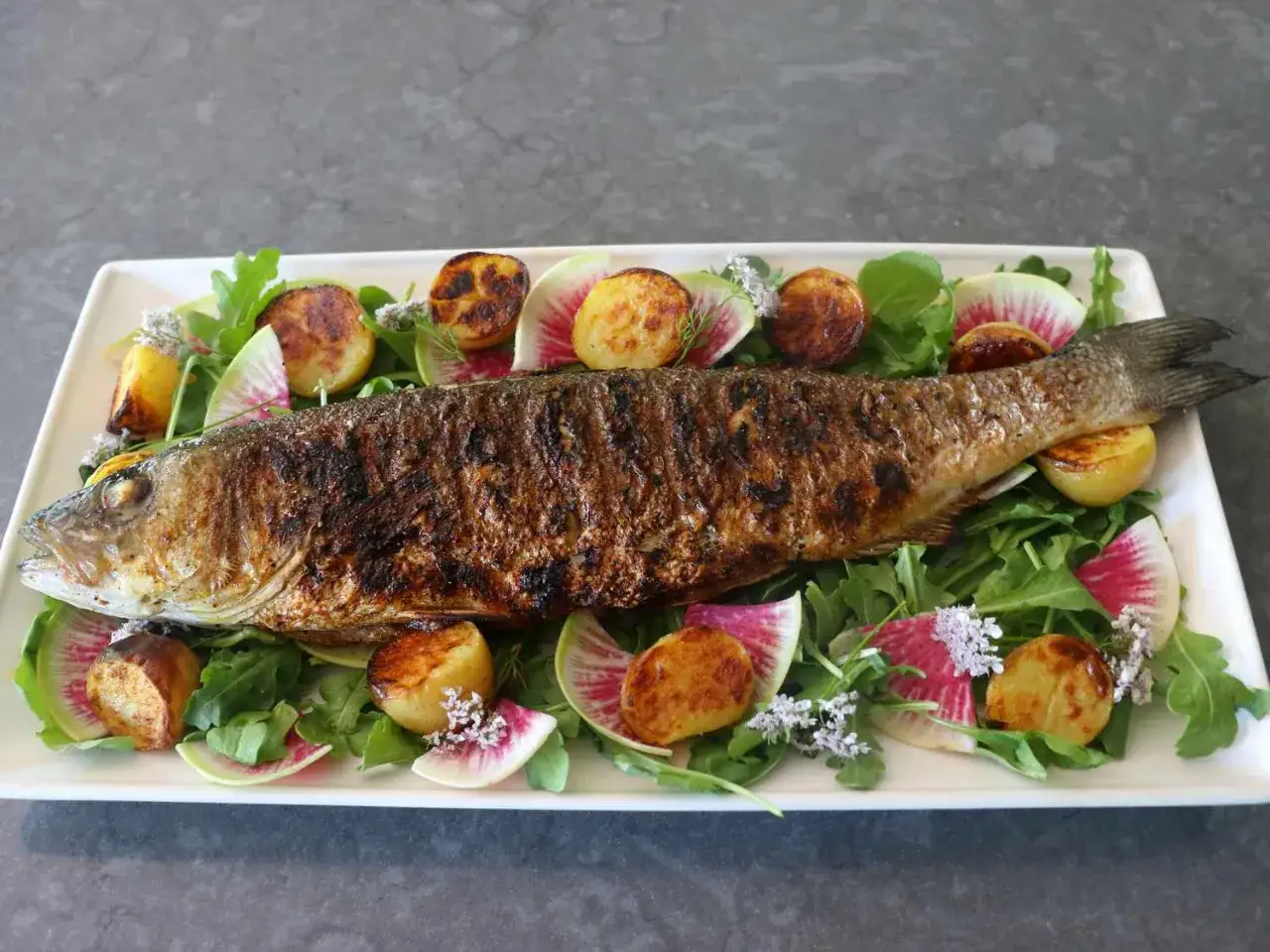 Bar au barbecue entier, servi sur un lit de roquette avec des pommes de terre rôties et des radis watermelon.