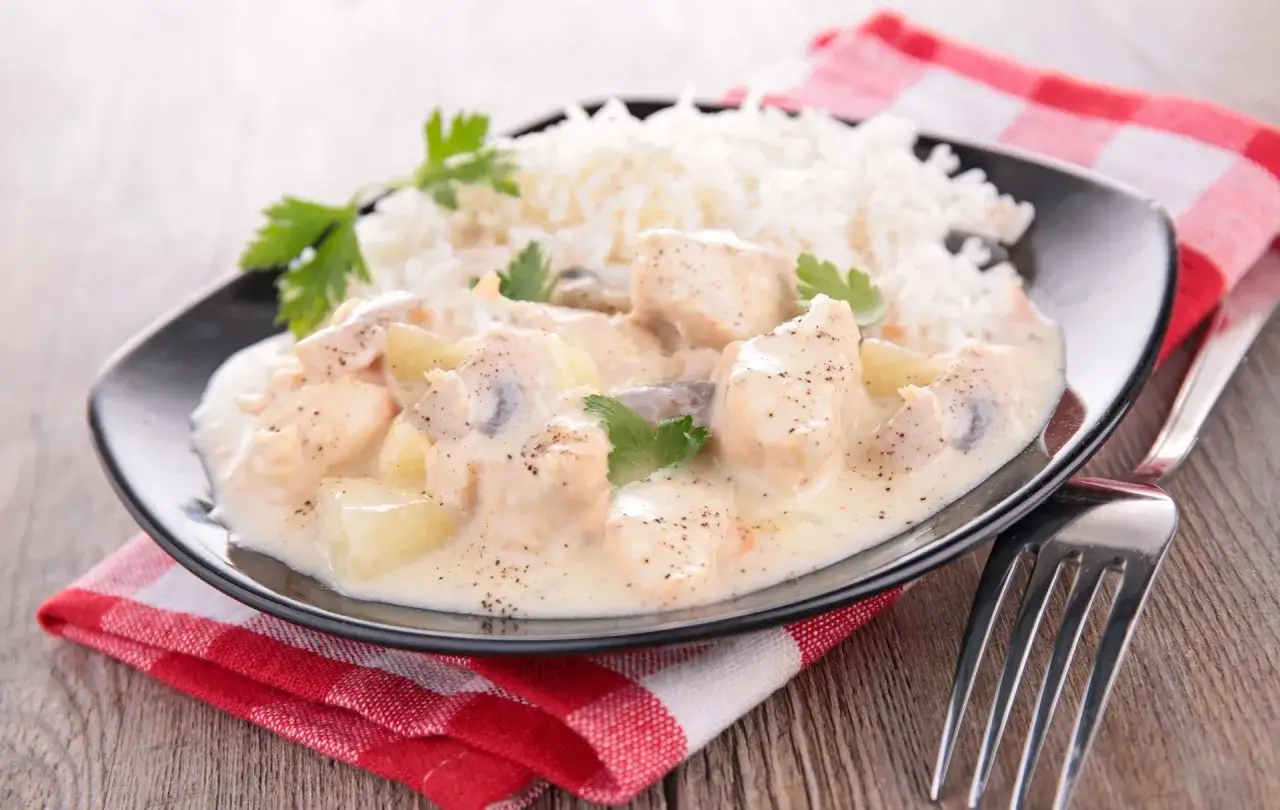 Une assiette de blanquette de volaille crémeuse avec des morceaux de poulet, des pommes de terre et des champignons, servie avec du riz blanc et garnie de persil frais.