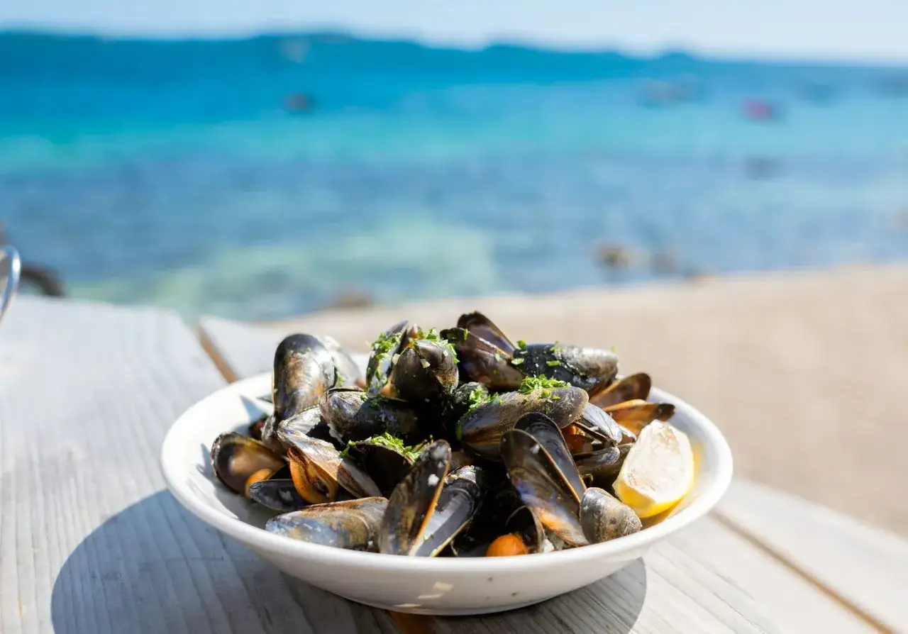 Un plat de moules savoureuses, garni de persil frais et d'un quartier de citron, sur une table en bois, avec la mer bleue en arrière-plan. Une recette moules parfaite pour l'été.