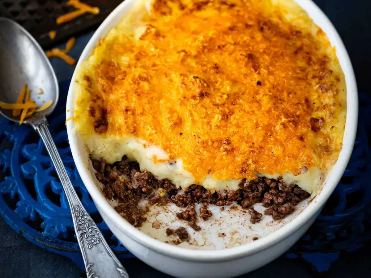 Un plat de parmentier de boeuf gratiné, avec une couche de purée onctueuse et une garniture de viande savoureuse. Une cuillère ornée est à côté.