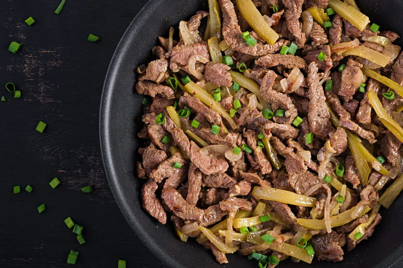 Boeuf aux oignons savoureux, garni de ciboule fraîche, dans une poêle sur fond sombre. Un plat réconfortant prêt à être dégusté.