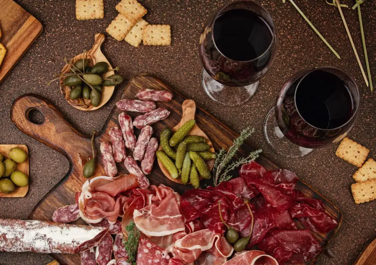 Plateau de charcuterie vari&eacute;e, cornichons, olives, c&acirc;pres et deux verres de vin rouge. Id&eacute;al pour savoir quel vin avec charcuterie.