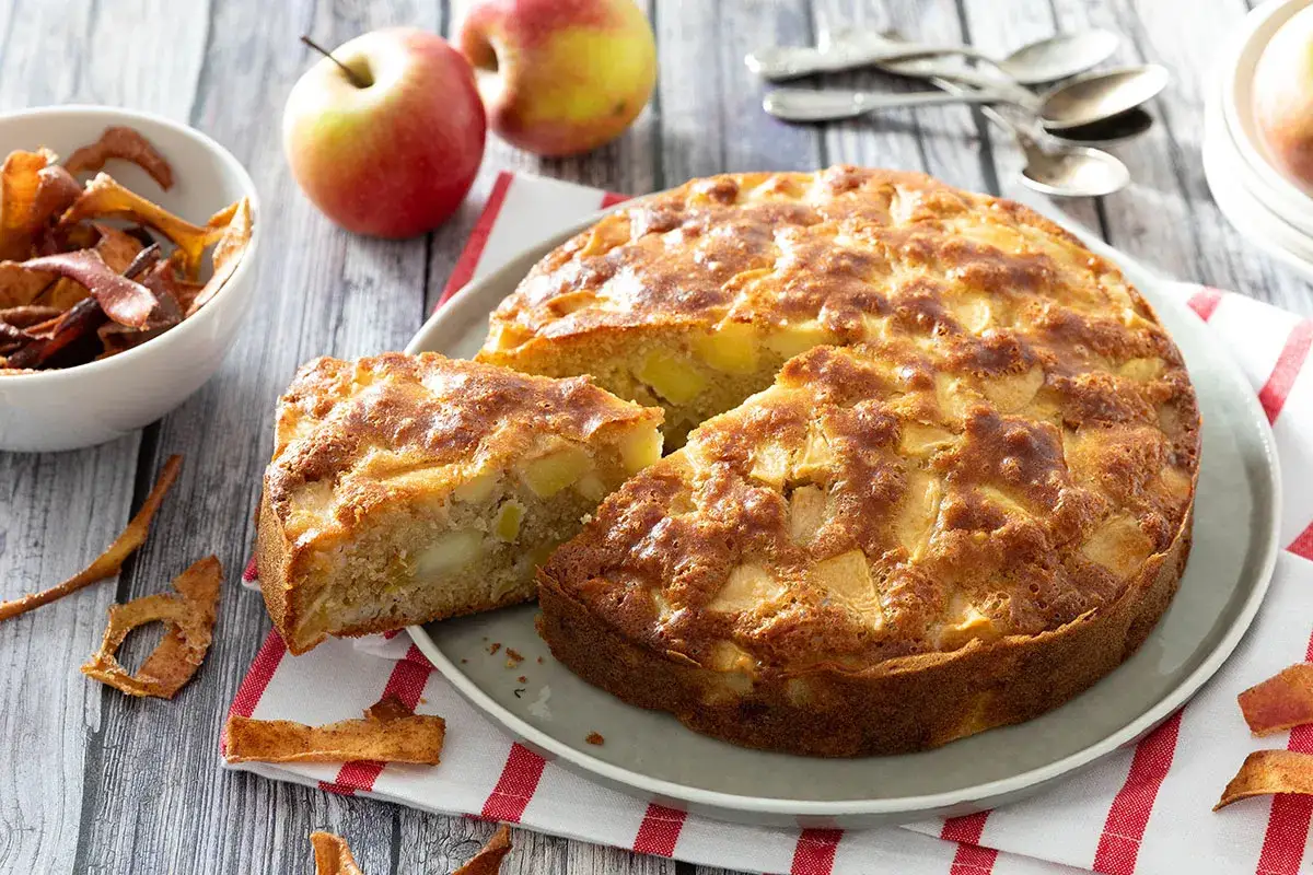 Un d&eacute;licieux moelleux aux pommes, dor&eacute; et garni de morceaux de fruits. Des pommes fra&icirc;ches et des chips de pommes sont dispos&eacute;s autour.
