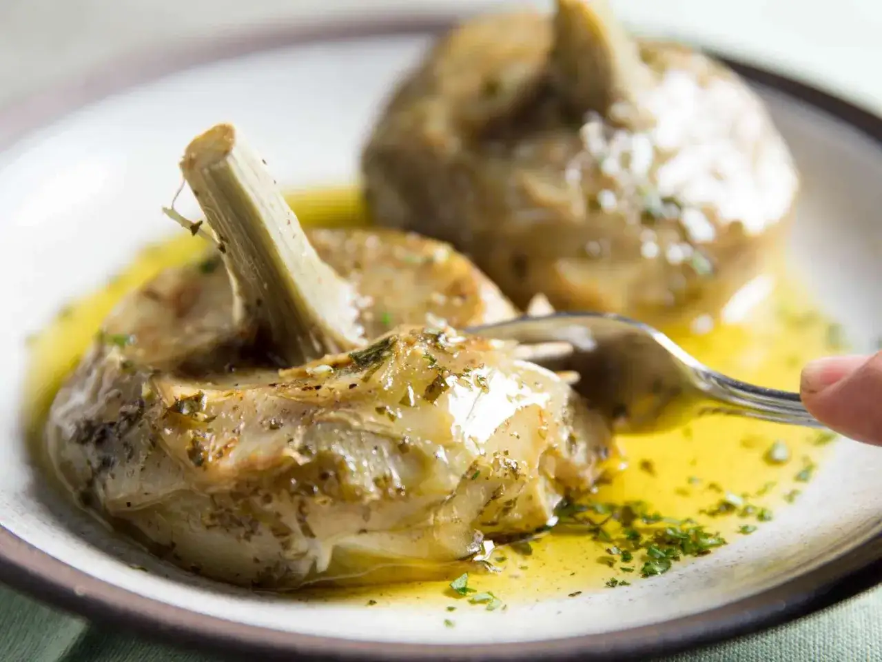 Artichauts à la romaine tendres, baignant dans une sauce dorée aux herbes, une fourchette prête à déguster ce plat savoureux.