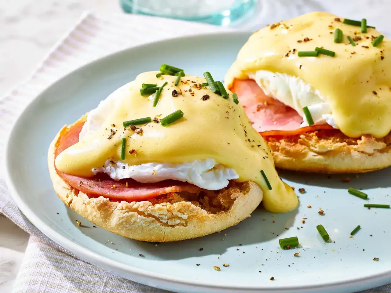 Deux œufs Bénédicte appétissants sur une assiette bleue, garnis de jambon, d'œuf poché et de sauce hollandaise, parsemés de ciboulette.