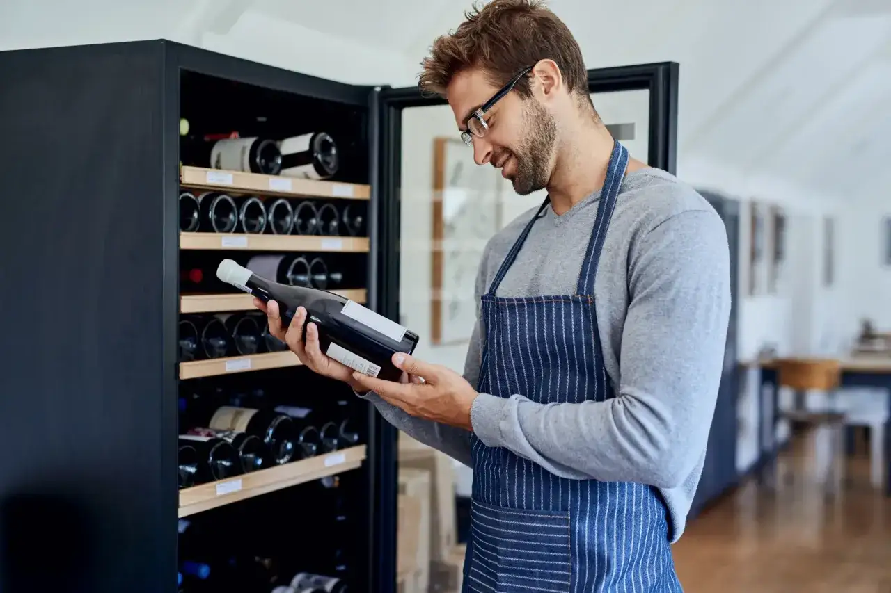 Un homme en tablier examine une bouteille de vin, entouré d'une cave à vins bien garnie.