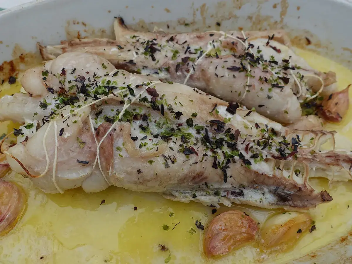 Deux morceaux de lotte au four, liés à la ficelle et parsemés d'herbes, baignent dans un jus doré avec des gousses d'ail rôties.
