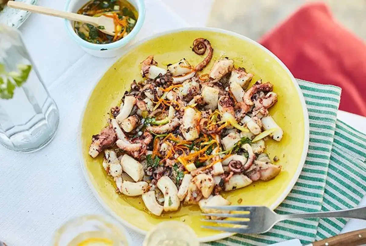 Délicieux poulpe à la plancha, servi sur une assiette jaune avec des herbes et des zestes d'agrumes. Un plat estival parfait.