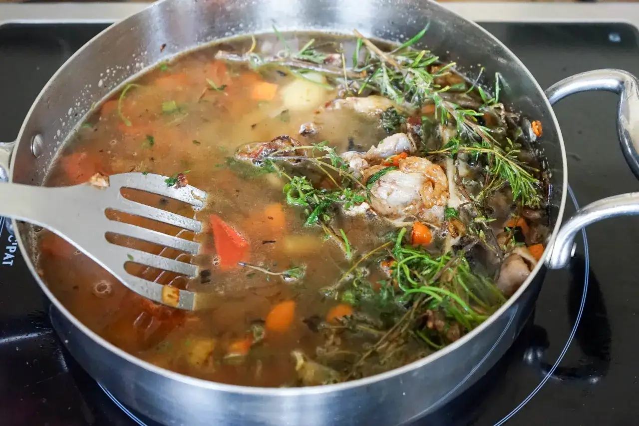 Un bouillon riche et savoureux mijote dans une casserole, avec des morceaux de viande, des carottes et des herbes aromatiques sur un fond brun.