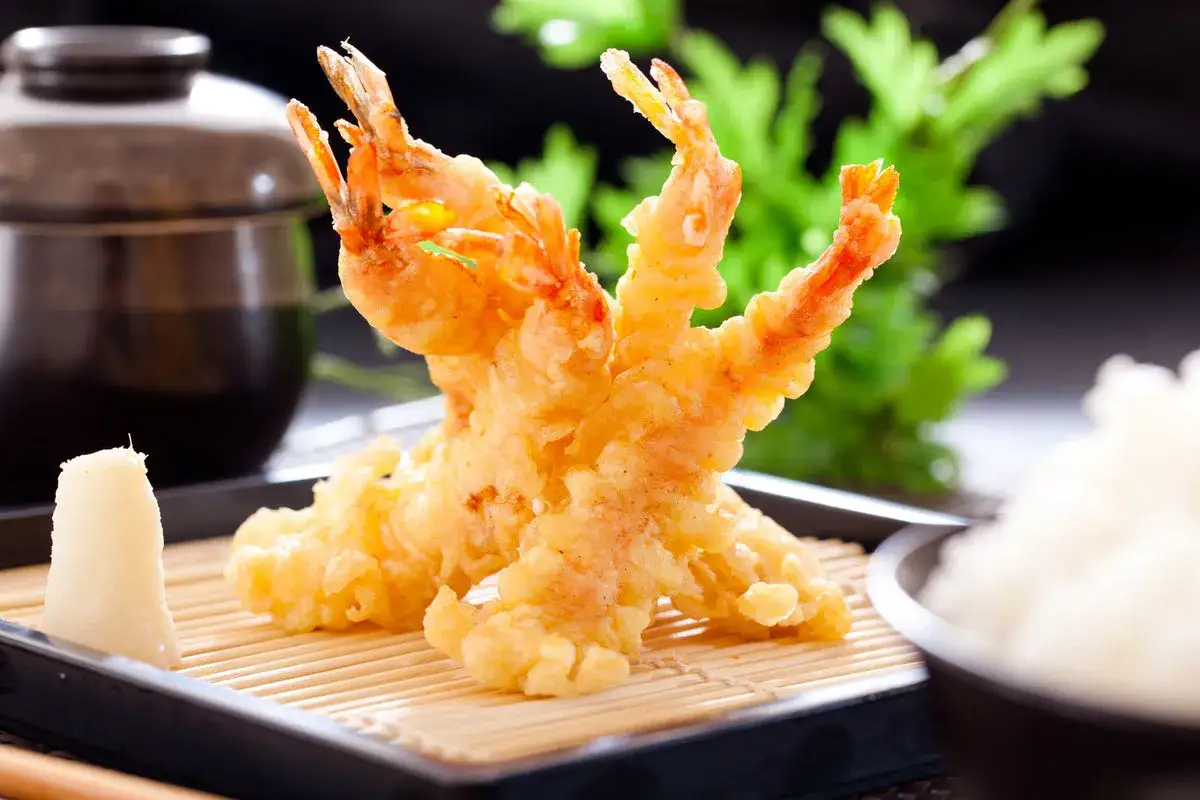 Trois beignets de crevette dorés et croustillants, servis sur un lit de bambou avec du riz blanc et une sauce foncée.