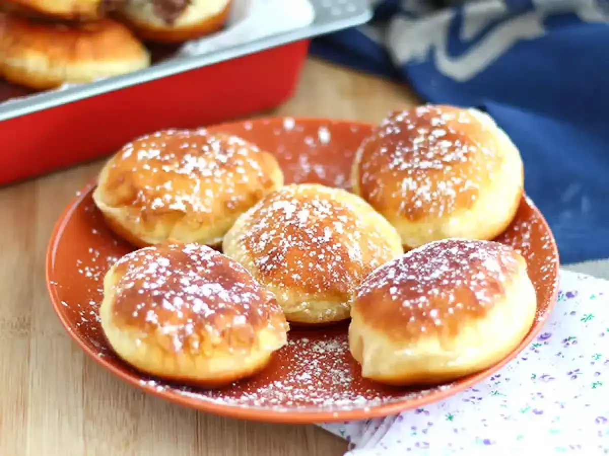 Cinq beignets au chocolat saupoudrés de sucre glace sur une assiette orange. Un beignet au chocolat est visible dans un plat rouge en arrière-plan.